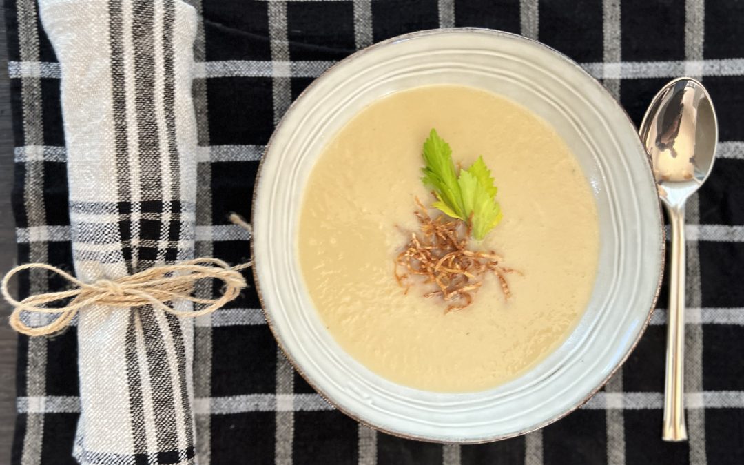 Selleriecremesuppe in einem weißen Suppenteller angerichtet mit Selleriestroh und Selleriegrün dekoriert, auf einem schwarz-weiß karierten Tischtuch mit einer Serviette und einem Löffel.