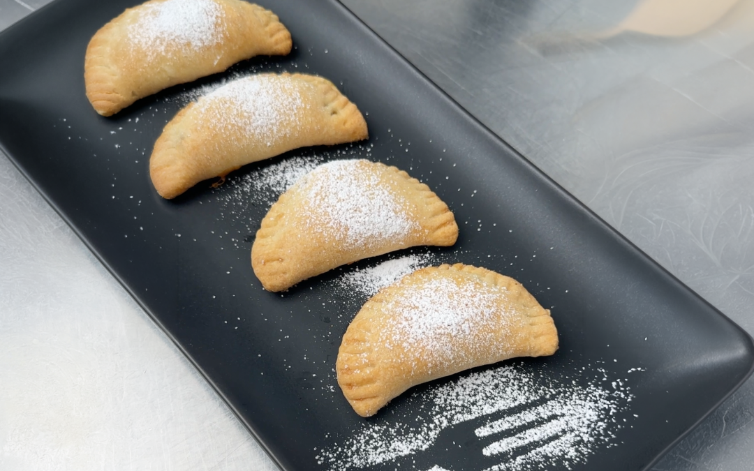 4 mit Staubzucker bestreute halbmondförmige Apfeltaschen auf einer schwarzen Servierplatte.