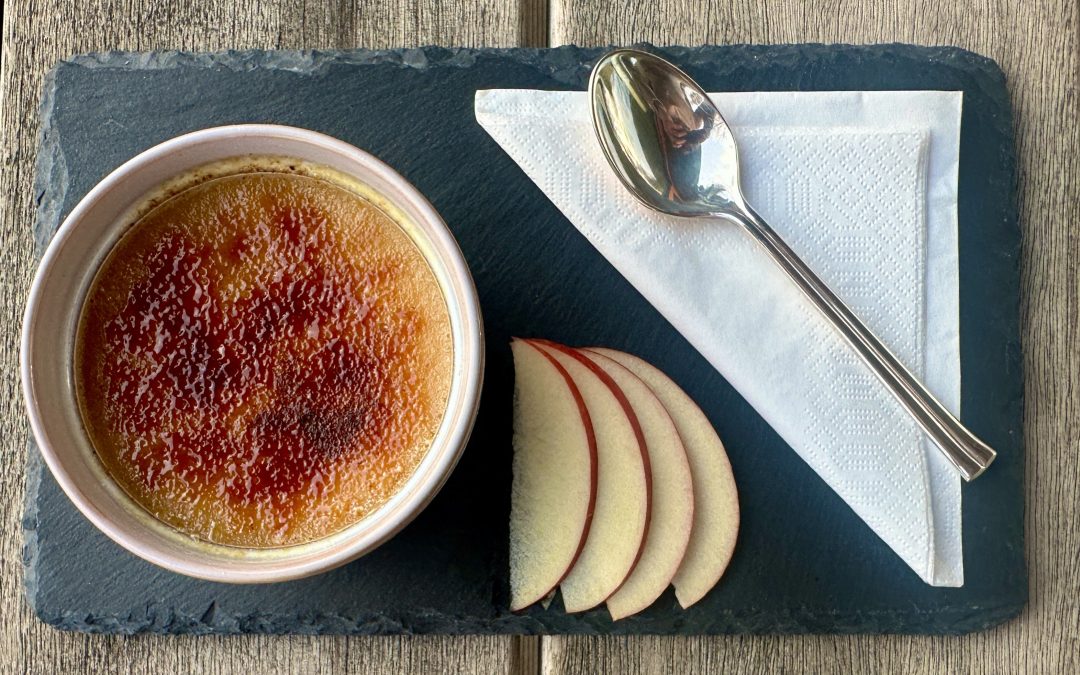 Bratapfel-Crème Brûlée auf einer schwarzen Schieferplatte mit einer weißen Serviette, kleinem Löffel und Apfelspalten-Garnitur auf einem Holztisch von oben fotografiert.